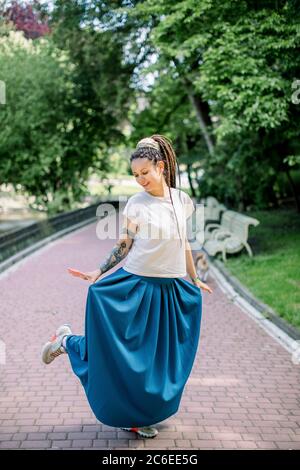 Portrait en longueur de jolie jeune fille avec des dreadlocks dansant dans le parc de la ville, regardant sa jupe longue bleue et souriante Banque D'Images