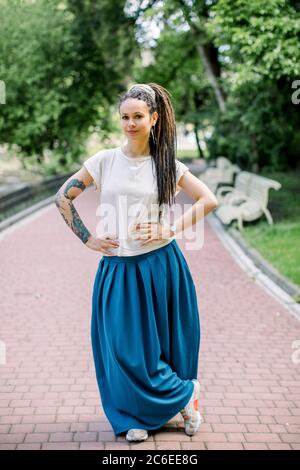 Portrait en longueur de fille hippster attrayante avec des dreadlocks et tatouages, portant une chemise blanche et une jupe bleue, posant à l'appareil photo dans un parc de la ville Banque D'Images