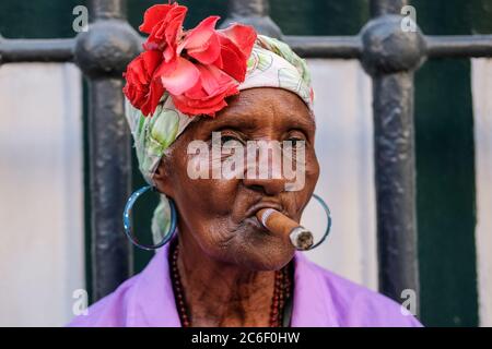 Une cubaine vêtue de couleurs fume un cigare alors qu'elle est assise sur le côté de la rue Habana Vieja à la Havane, Cuba. Banque D'Images