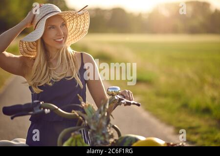 Belle femme portant un chapeau de paille à large bord shopping pour des produits frais dans le pays en faisant rouler sa bicyclette le long d'une voie rurale dans un pays chaud d'evenin Banque D'Images