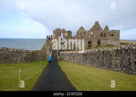 Lady en manteau bleu clair descend le chemin d'entrée vers les ruines spectaculaires du château de Dunluce près de Portrush, Co. Antrim, Irlande du Nord Banque D'Images