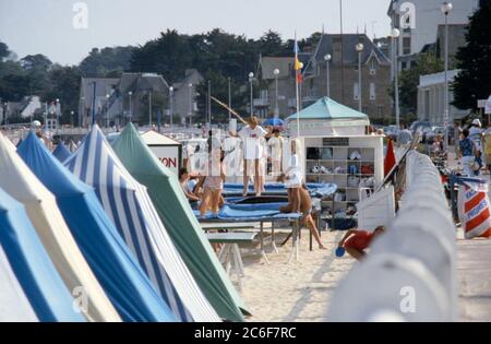 Les enfants jouant et sautant sur les trampolines dans les clubs de la plage, et les gens marchant le long de la promenade traditionnelle au bord de la mer à St Cast le Guildo, Bretagne, France 1984. Banque D'Images