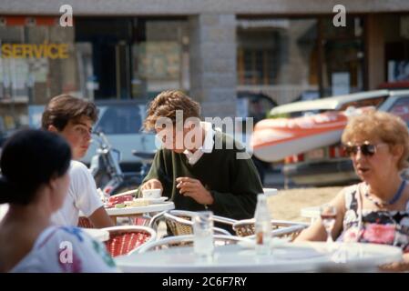 Personnes assises à des tables à l'extérieur, en dégustant des boissons et de la nourriture dans un café de la place à St Cast le Guildo, Bretagne, France 1984. Banque D'Images