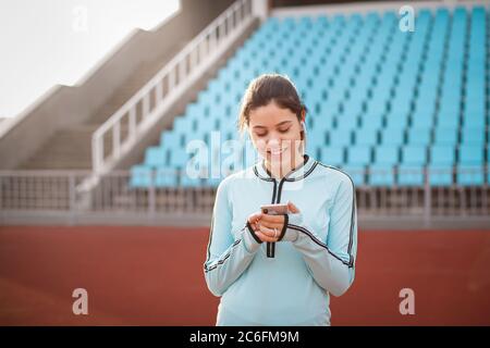 Belle fille regarde dans le téléphone sur le fond du stade au coucher du soleil. La fille dans un uniforme de sport bleu sourit après un entraînement. Banque D'Images