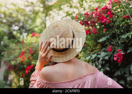 Chapeau de soleil paille d'été. Fille marchant par des roses en fleur dans le jardin. Accessoires tendance. La femme aime la nature Banque D'Images