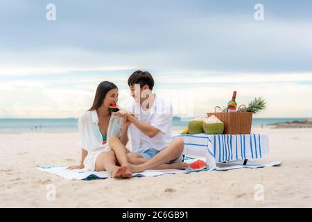 Jeune couple asiatique assis sur la couverture de pique-nique et mangeant de la pastèque sur la plage et près de la mer avec des fruits tropicaux en arrière-plan. Été, vacances, Banque D'Images