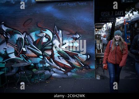 Photographie de rue.Une femme portant un manteau rouge marchant devant une murale de graffiti d'art de rue à Sydney, en Australie. Banque D'Images