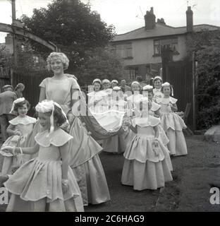 Années 1950, historique, avec un grand sourire sur son visage, la Reine Rose nouvellement couronnée de la ville, Farnworth, Bolton, Lancashire, Angleterre, Royaume-Uni avec son entourage de jeunes filles tenant le train, marchant dans une procession à travers le parc local. Remontant aux années 1880, le festival annuel Rose Queen, organisé en juin, est devenu un événement annuel majeur dans de nombreuses villes et villages du Royaume-Uni, en particulier dans le Lancashire, connu sous le nom de comté de la rose rouge, après les guerres des Roses de 1455-87. Banque D'Images