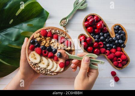 Smoothie Acai, granola, graines, fruits frais dans un bol en bois aux mains des femmes avec une cuillère de cactus. Assiette remplie de baies sur fond de bois blanc Banque D'Images