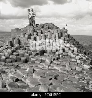 Dans les années 1950, deux visiteurs masculins historiques portant des vestes et des cravates de sport, se tenant sur les rochers hexagonaux de la côte, à la célèbre chaussée des géants, à Co. Antrim, en Irlande du Nord. Les roches sont une série de colonnes de basalte emboîtées, le résultat d'une éruption volcanique ancienne de la fissure. Banque D'Images