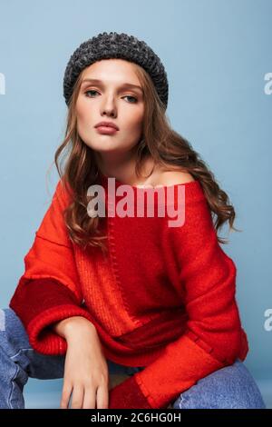 Jeune belle femme pensive avec cheveux ondulés dans le chapeau et chandail rouge regardant soigneusement dans l'appareil photo assis sur fond bleu Banque D'Images