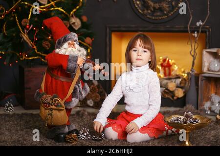 Petite fille assise sur le sol dans de belles décorations de Noël. Fille jouant avec un jouet Santa Claus. Préparation de la nouvelle année. Banque D'Images