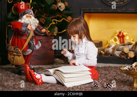 Petite fille assise sur le sol dans de belles décorations de Noël. Fille lisant un jouet de conte de fées Santa Claus. Fille jouant avec un jouet Santa Claus. Banque D'Images