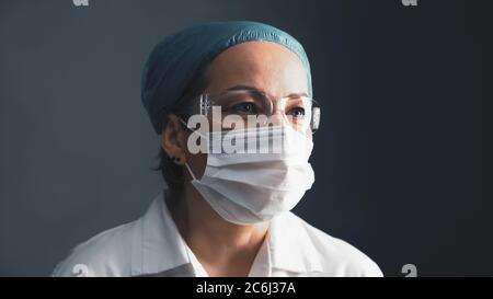 Médecin masqué dans les lunettes de protection regardant sur le côté. Femme de race blanche d'âge moyen, en blanc, sur fond gris foncé. Gros plan portrait. Santé Banque D'Images