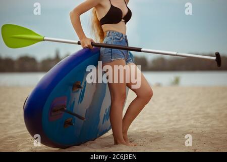 Jeune femme attrayante debout à côté du paddle board, SUP. Vie active, sport, loisirs concept. Femme caucasienne à bord en été soir. Vacances, station, plaisir. Rogné. Banque D'Images