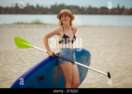 Jeune femme attrayante debout à côté du paddle board, SUP. Vie active, sport, loisirs concept. Femme caucasienne à bord en été soir. Vacances, station, plaisir. Banque D'Images