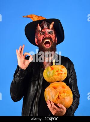 Spooky Halloween party concept. Avec les cornes du démon et du mal sourire détient jack o lanternes et montre ok signe. Monster avec décorations d'octobre. Dans l'homme witchers hat détient les citrouilles sur fond bleu Banque D'Images