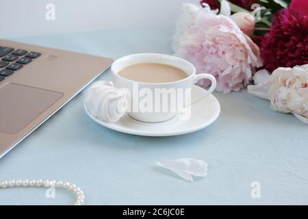 Espace de travail féminin avec ordinateur portable, tasse de café, collier de perles et fleurs de pivoine sur fond bleu. Femme dans le concept d'affaires. Flat lay, vue de dessus. Banque D'Images