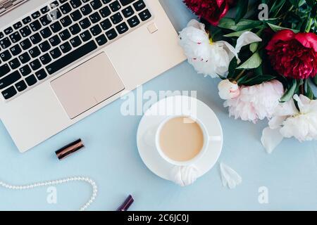 Espace de travail féminin avec ordinateur portable, tasse de café, rouge à lèvres, collier de perles et fleurs de pivoine sur fond bleu. Femme dans le concept d'affaires. Flat lay, vue de dessus. Banque D'Images