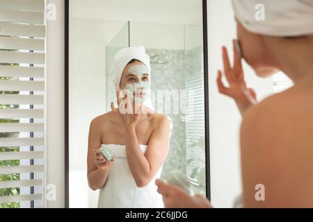 Femme appliquant le paquet de visage en regardant dans le miroir Banque D'Images