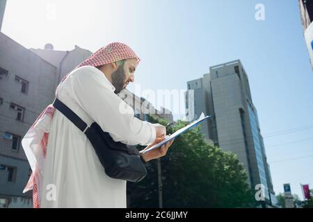 Contrat. Riche portrait d'homme arabe lors de l'achat de biens immobiliers, centre d'affaires en ville. Ethnicité, culture. Homme confiant dans les vêtements traditionnels faisant une affaire réussie. Finances, économie. Banque D'Images