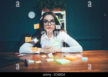 Femme folle avec des étiquettes de rappel assise au bureau. Beaucoup d'autocollants de mémoire collés sur jeune femme d'affaires caucasienne. Le multitâche. Date limite Banque D'Images