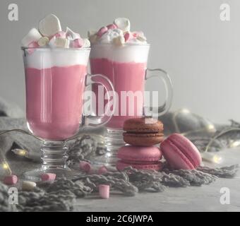 Chocolat chaud rose avec crème fouettée et bonbons guimauve dans 2 tasses en verre, macarons français doux pastel et lumières. Banque D'Images