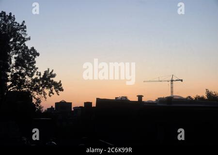 Vue sur l'horizon de l'aube avec une grue de levage en arrière-plan et au premier plan des toits de la maison et des branches d'arbres Banque D'Images