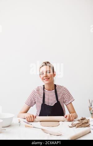 Jolie fille souriante dans un tablier noir et un t-shirt rayé assis à la table avec une goupille et travaillant avec de l'argile, regardant joyeusement dans la caméra à pott Banque D'Images
