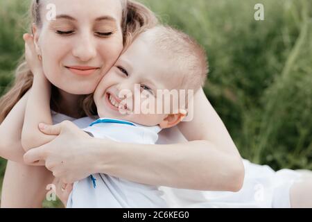 Petit garçon souriant embrassant sa mère heureuse qui est assise avec les yeux fermés dans la nature. Mère et fils. Bonne famille Banque D'Images