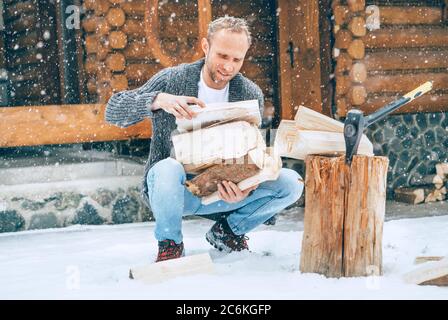 Homme collectant du bois de chauffage haché sur cour enneigée pour une cheminée avec fond de flocons de neige lourds. Image concept vacances campagne d'hiver Banque D'Images