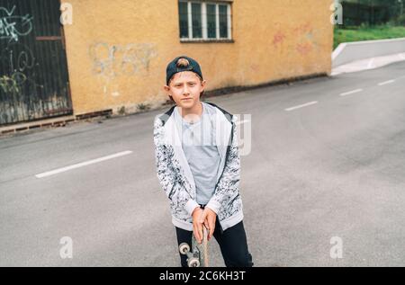 Jeune skateboarder garçon dans un portrait de rue de casquette de baseball tenant le skateboard sur la route d'asphalte près d'un mur de graffiti de grunge. Génération de jeunes temps libre sp Banque D'Images