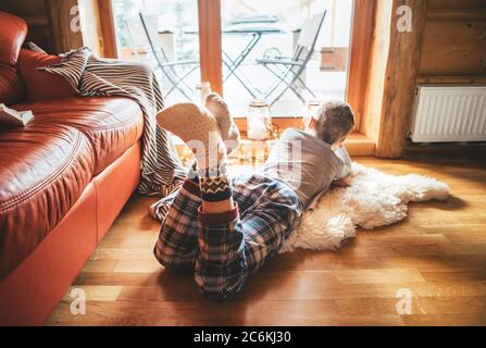 Garçon couché sur le sol sur une peau de mouton et regardant dans la fenêtre, habillé dans un confortable pyjama maison et chaussettes chaudes Banque D'Images