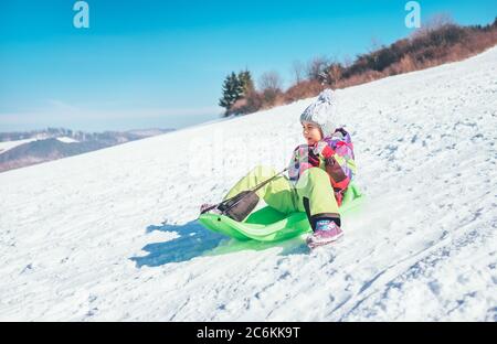 Bonne rire petite fille glissant vers le bas de la pente de neige. Image de concept de dépenses de vacances d'hiver drôle. Banque D'Images