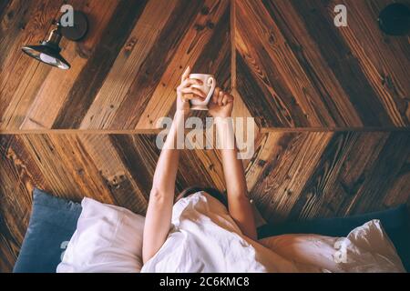 Paresseuse femme couchée sous la couverture blanche sur le lit en lin et tenant la tasse de café frais tôt le matin. Une journée de repos et un café au lit Banque D'Images