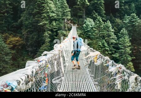 Jeune photographe souriante traversant le canyon au-dessus du pont suspendu décoré de drapeaux de prière tibétains multicolores sur les gorges. Everest B. Banque D'Images