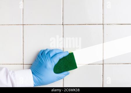 une main dans un gant en caoutchouc bleu contient une éponge de nettoyage en microfibre jaune-vert, en lavant un mur de carreaux blancs Banque D'Images