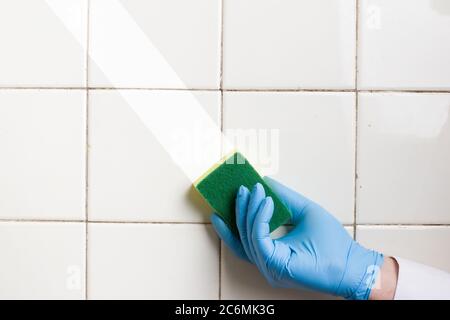 une main dans un gant en caoutchouc bleu contient une éponge de nettoyage en microfibre jaune-vert, en lavant un mur de carreaux blancs Banque D'Images