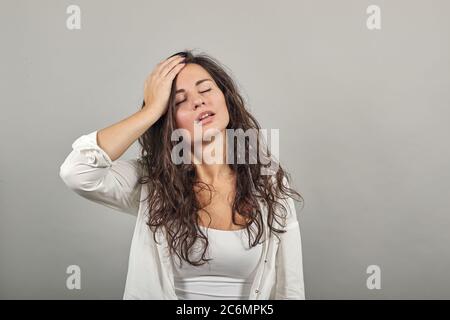 Stress ou mal de tête grimaquant dans la douleur maintient l'arrière du cou indiquant l'emplacement. Fatigue pendant le travail de travail. Jeune femme attrayante, blouse blanche habillée, avec des yeux bruns, cheveux bouclés, gris Banque D'Images