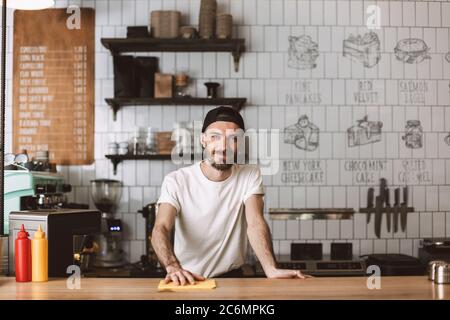 Le barista souriant se tient debout dans le chapeau et le comptoir de la barre d'essuyage tout en regardant avec plaisir dans l'appareil photo du café. Banque D'Images