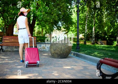 Femme en chapeau de paille et masque de protection rose, marche dans la rue avec une valise Banque D'Images