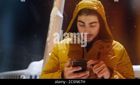 Jeune homme dans un message à capuche avec le téléphone dans sa main sur une rue enneigée de nuit Banque D'Images