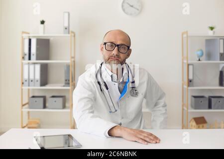 Un médecin mature qui regarde la caméra écoute le patient à l'aide d'une conférence avec webcam, assis à une table dans le bureau de la clinique. Banque D'Images