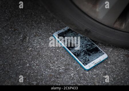 L'écran du smartphone est fissuré à plat sur la route, car le déplacement d'un pneu de voiture à l'avant et à l'arrière-plan est flou Banque D'Images