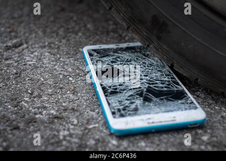 L'écran du smartphone est fissuré à plat sur la route, car le déplacement d'un pneu de voiture à l'avant et à l'arrière-plan est flou Banque D'Images