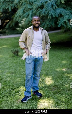 Portrait d'été en plein air d'un homme africain souriant en Jean, t-shirt blanc et maillot beige décontracté regardant l'appareil photo tout en se tenant debout sur l'herbe verte Banque D'Images