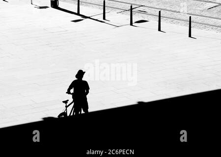 Silhouette d'un homme solitaire qui marche de l'ombre à côté de son vélo tout en traversant la place de la ville un jour chaud d'été, vue arrière à angle élevé Banque D'Images