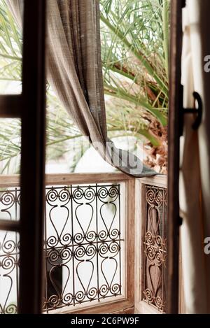 Vue à travers les portes-fenêtres sur un balcon orné avec des rideaux en cascade et un palmier en arrière-plan à Marrakech, au Maroc. Banque D'Images