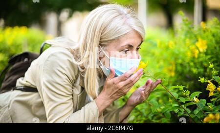 Femme essayant de sentir une fleur tout en portant un masque dans le parc, concept de pandémie de coronavirus Banque D'Images