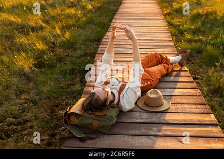 Une femme naturaliste de combinaison d'orange fait le selfie sur le téléphone, reposant sur un sac à dos sur un chemin en bois à travers le marais de tourbière dans le parc national de la faune Banque D'Images